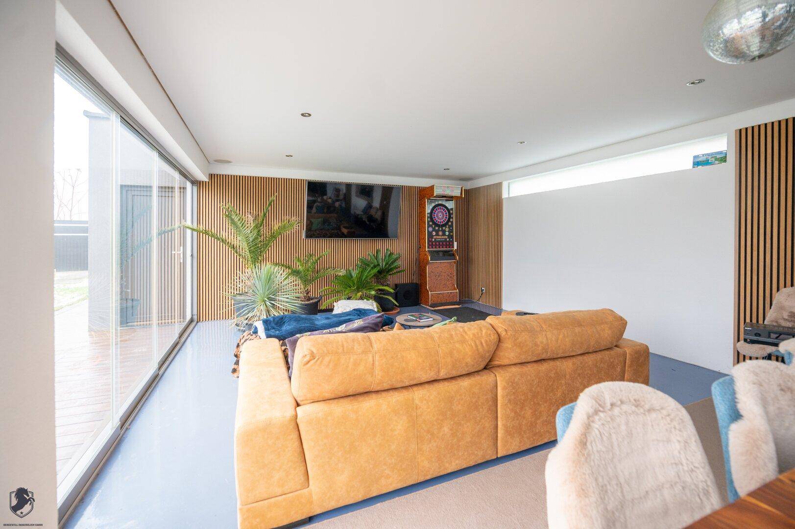 Gemütliches Wohnzimmer mit großem Sofa, Medienwand aus Holzlamellen und bodentiefen Fenstern zum Garten.