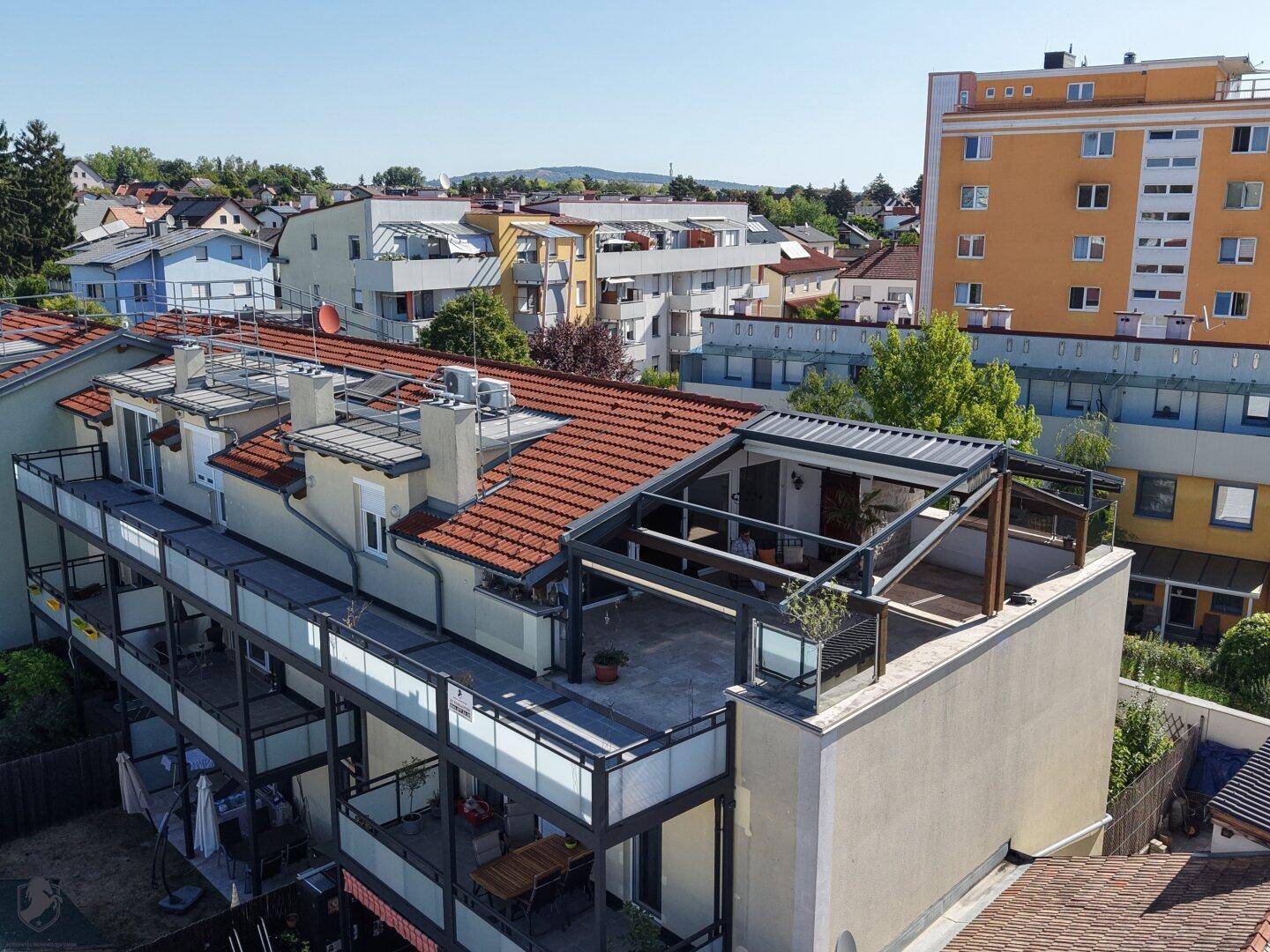 Luftaufnahme des Gebäudes mit Dachterrasse und umliegender Wohnbebauung unter blauem Himmel.