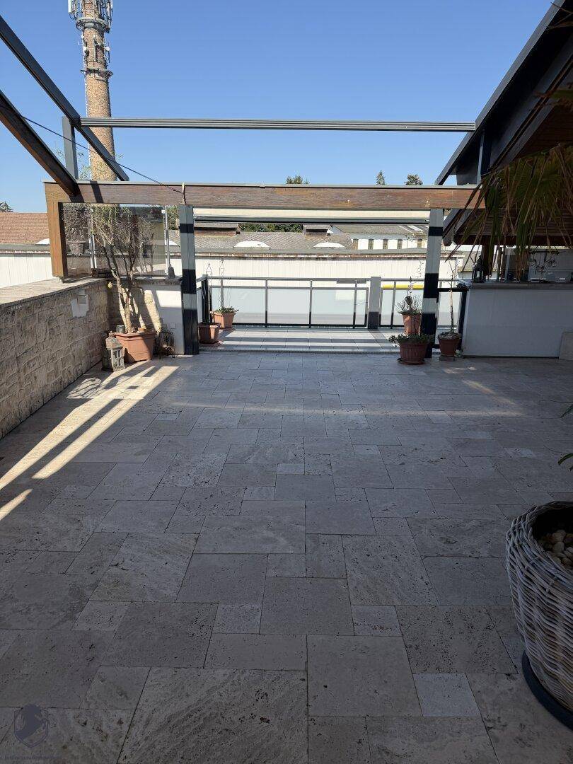 Weitläufige Dachterrasse mit hellem Fliesenboden und offener Pergola-Struktur unter blauem Himmel.