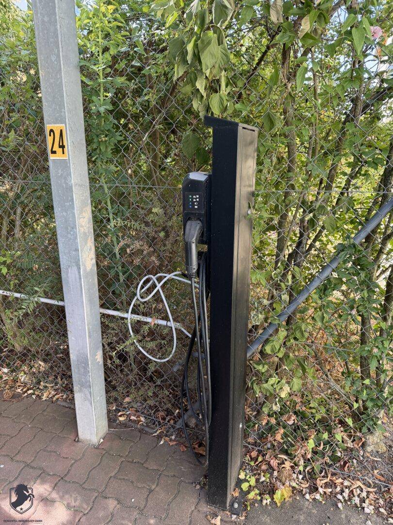 Ladestation für Elektrofahrzeuge im Freien, umgeben von Grün und einem Zaun.