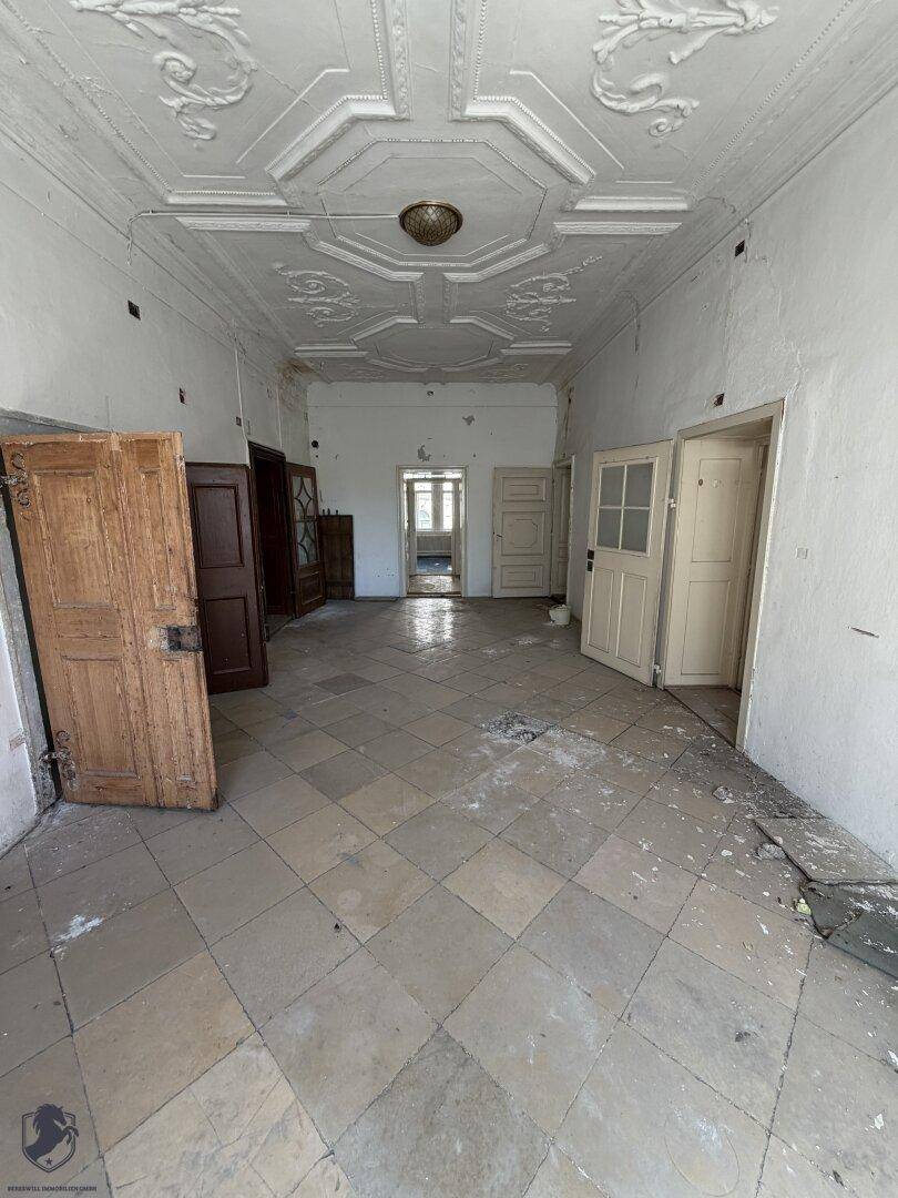 Großer leerer Raum mit Fliesenboden, Stuckdecke und mehreren Türen, bereit zur Renovierung.