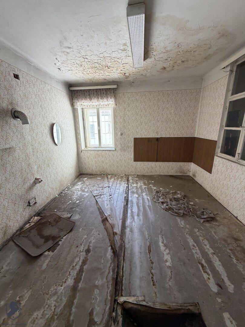 Kleiner Raum mit abgenutztem Holzboden, Fenster und Spuren von Wandinstallationen, renovierungsbedürftig.