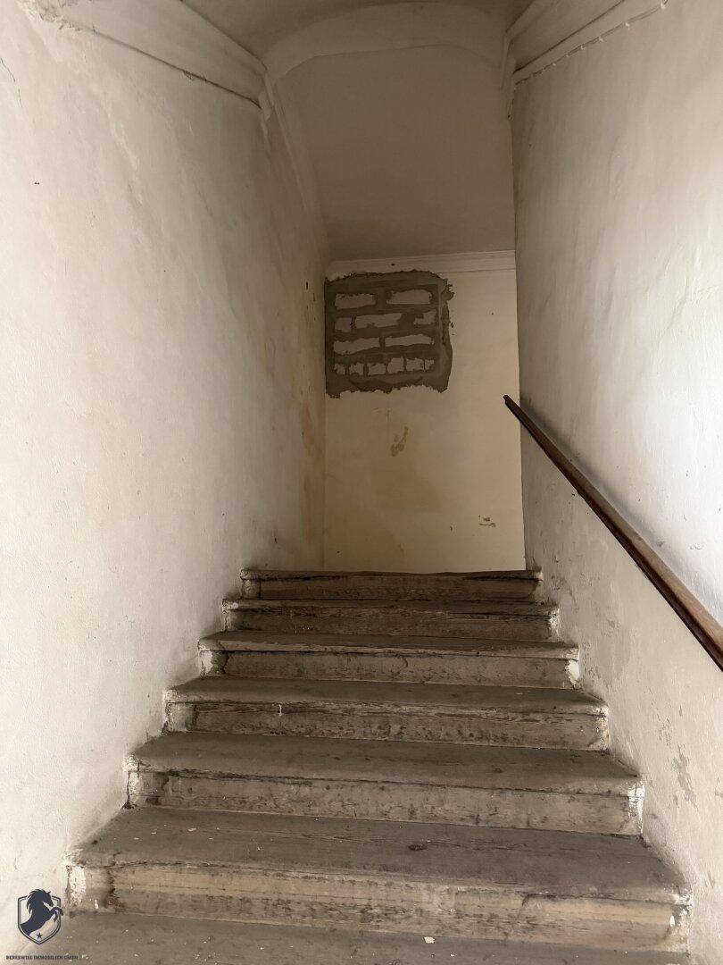 Alter Treppenaufgang mit einfacher Holzhandlauf und sichtbaren Mauerresten in der Wand.