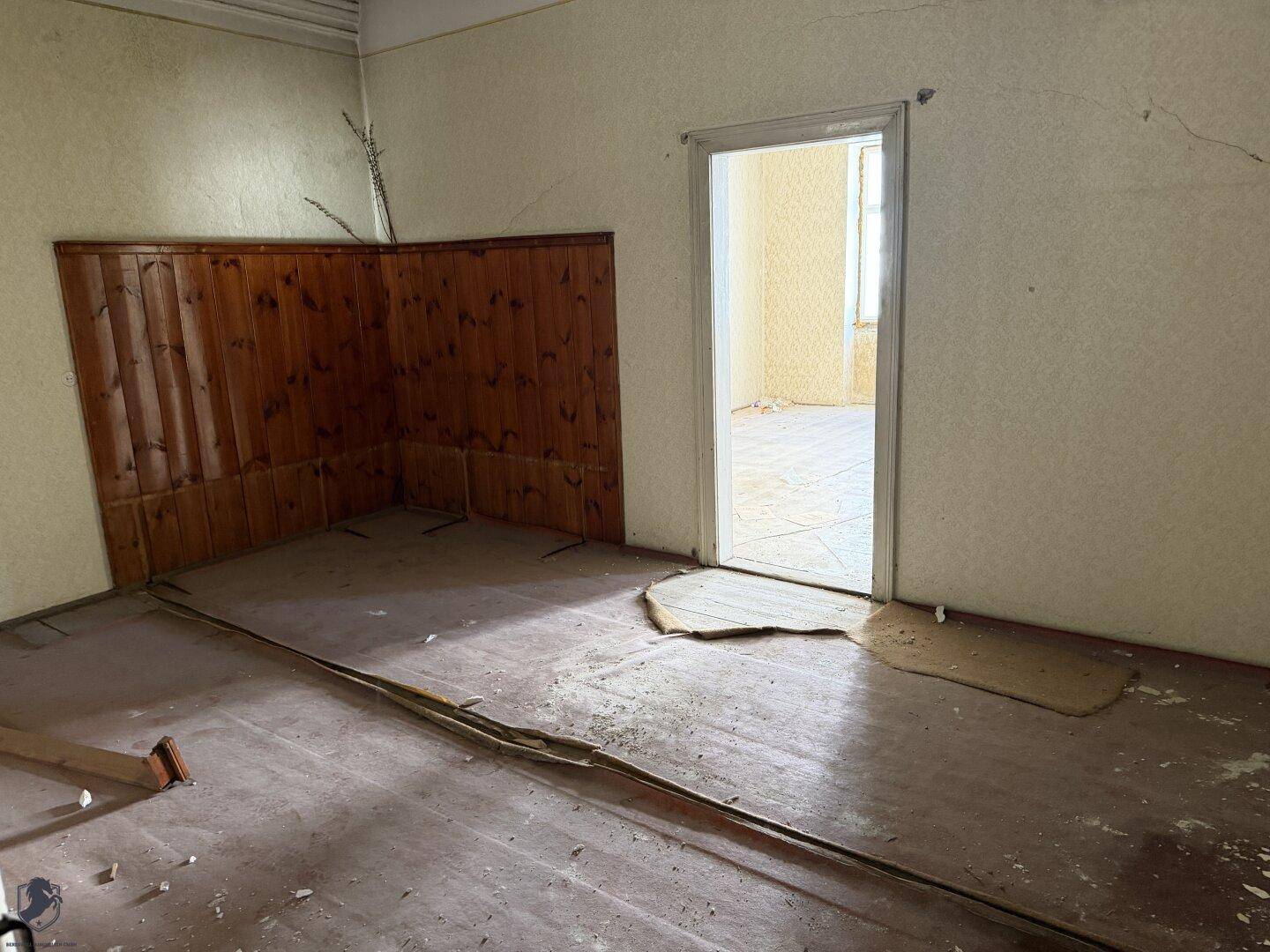 Leerer Raum mit Holzboden, Holzvertäfelung an einer Wand und Blick in einen weiteren Raum.