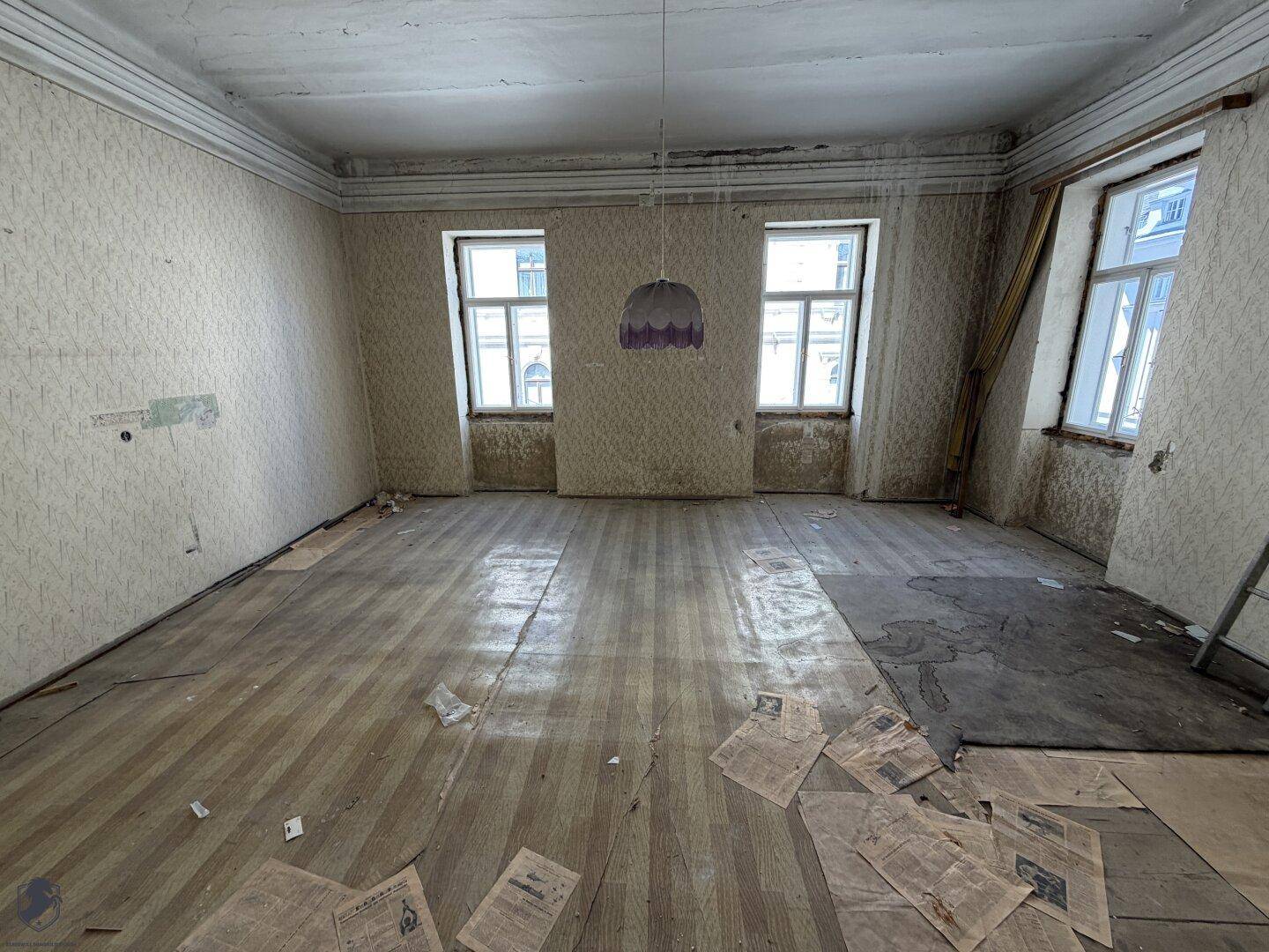Geräumiger, leerer Raum mit Holzboden, zwei Fenstern und beschädigter Tapete, renovierungsbedürftig.