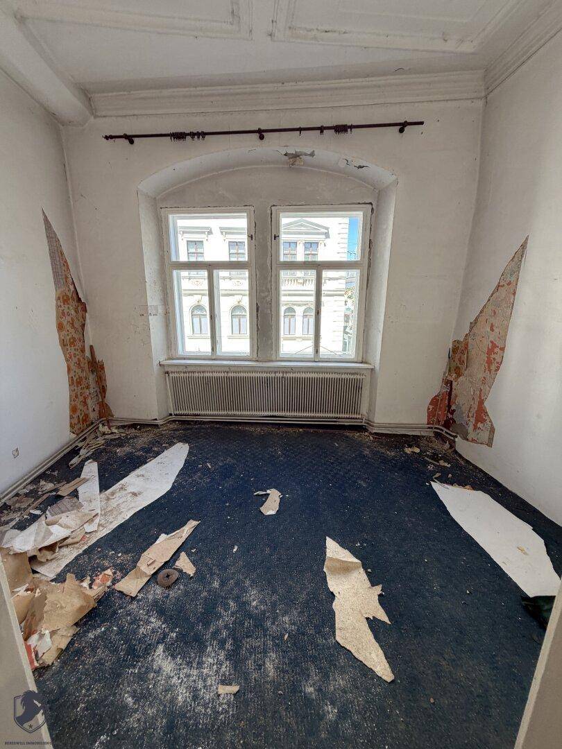 Leerer Raum mit Teppichboden, zwei Fenstern und abblätternder Wandfarbe, renovierungsbedürftig.