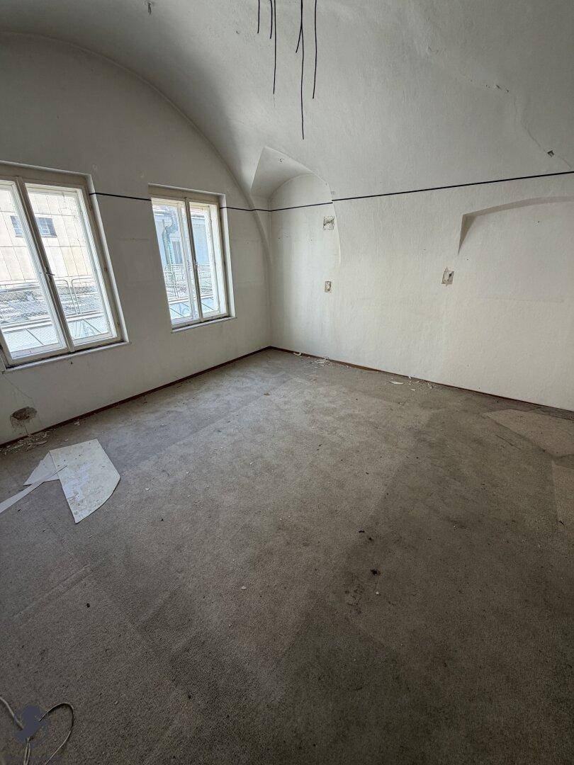 Leerer Raum mit Teppichboden, schrägen Wänden und zwei Fenstern, die Licht hereinlassen.