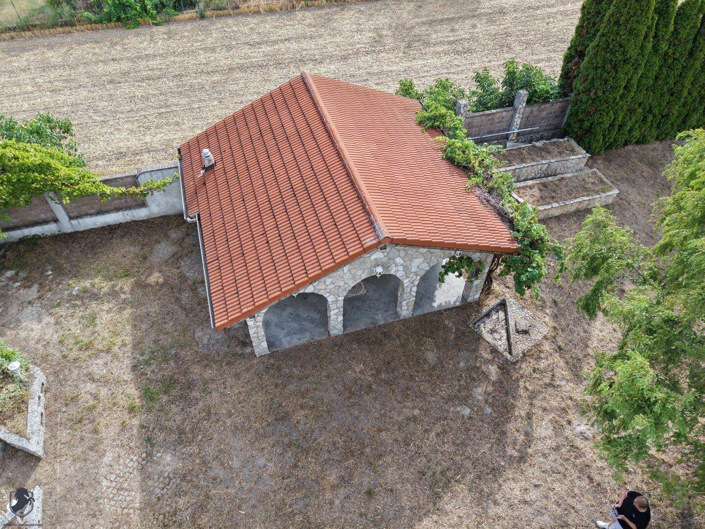 Luftaufnahme eines Gartenpavillons mit Ziegeldach und Steinbögen, umgeben von trockenem Gras.