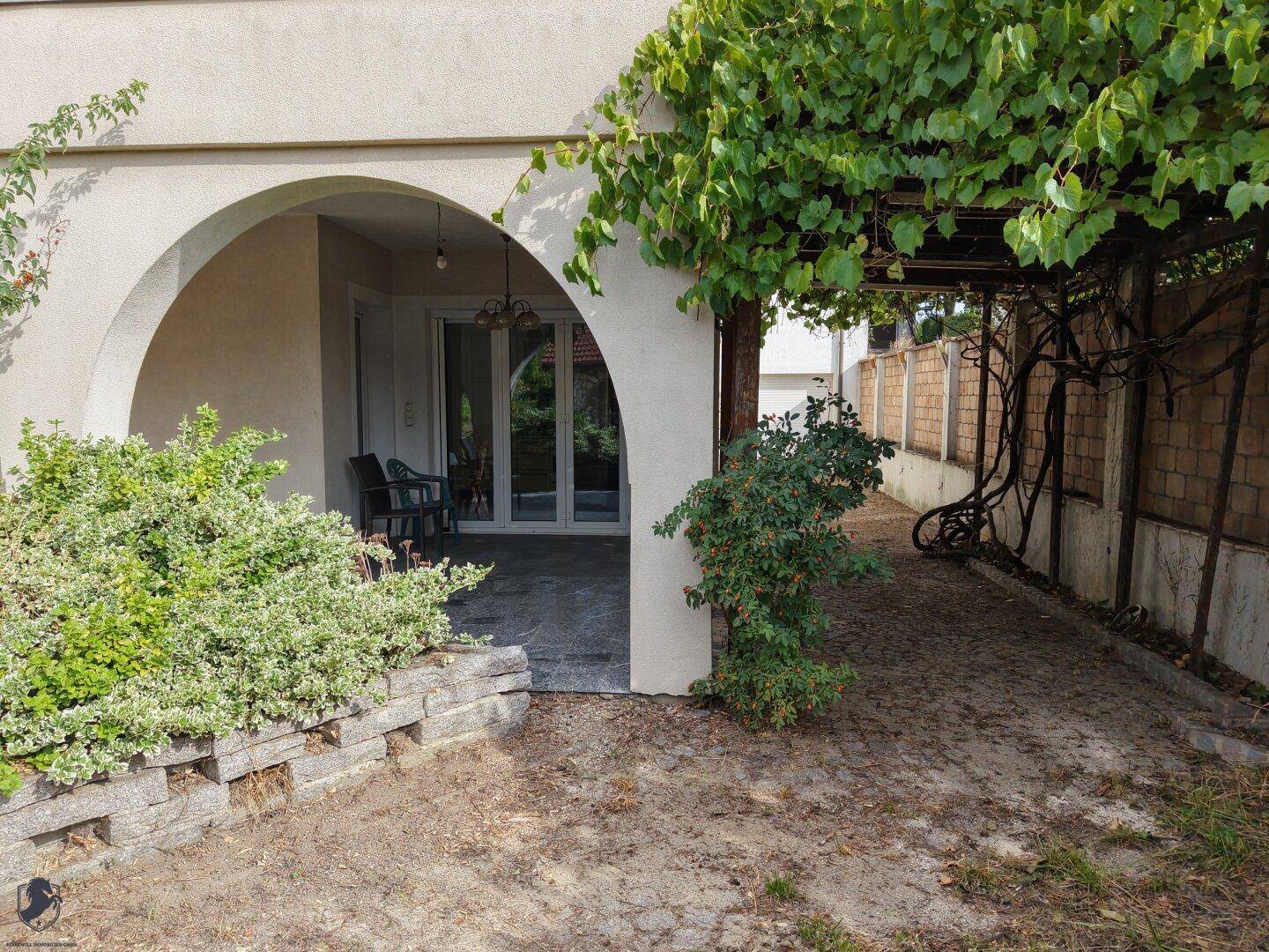 Überdachte Loggia mit Sitzgelegenheiten und Zugang zum Innenbereich, umgeben von Grünpflanzen.