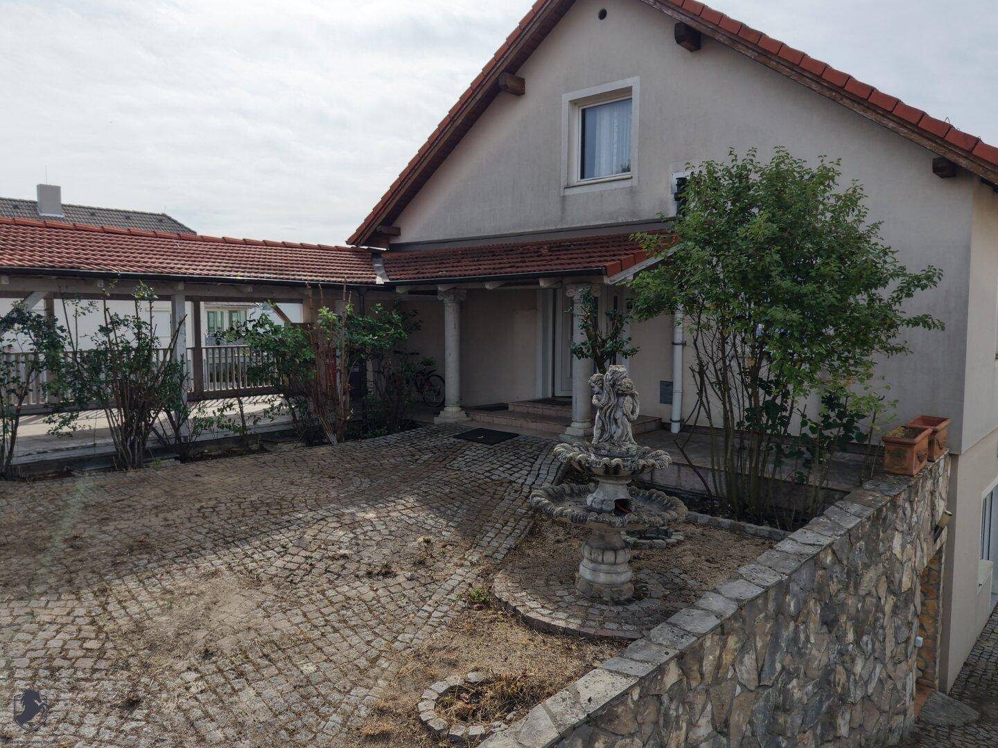 Eingangsbereich des Hauses mit Säulen, gepflastertem Vorplatz und einem dekorativen Brunnen.