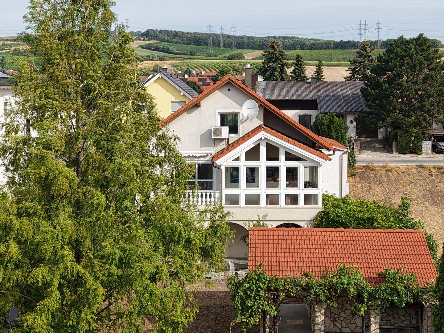 Hausansicht mit Wintergarten und Balkon, umgeben von grüner Landschaft und Weinbergen.