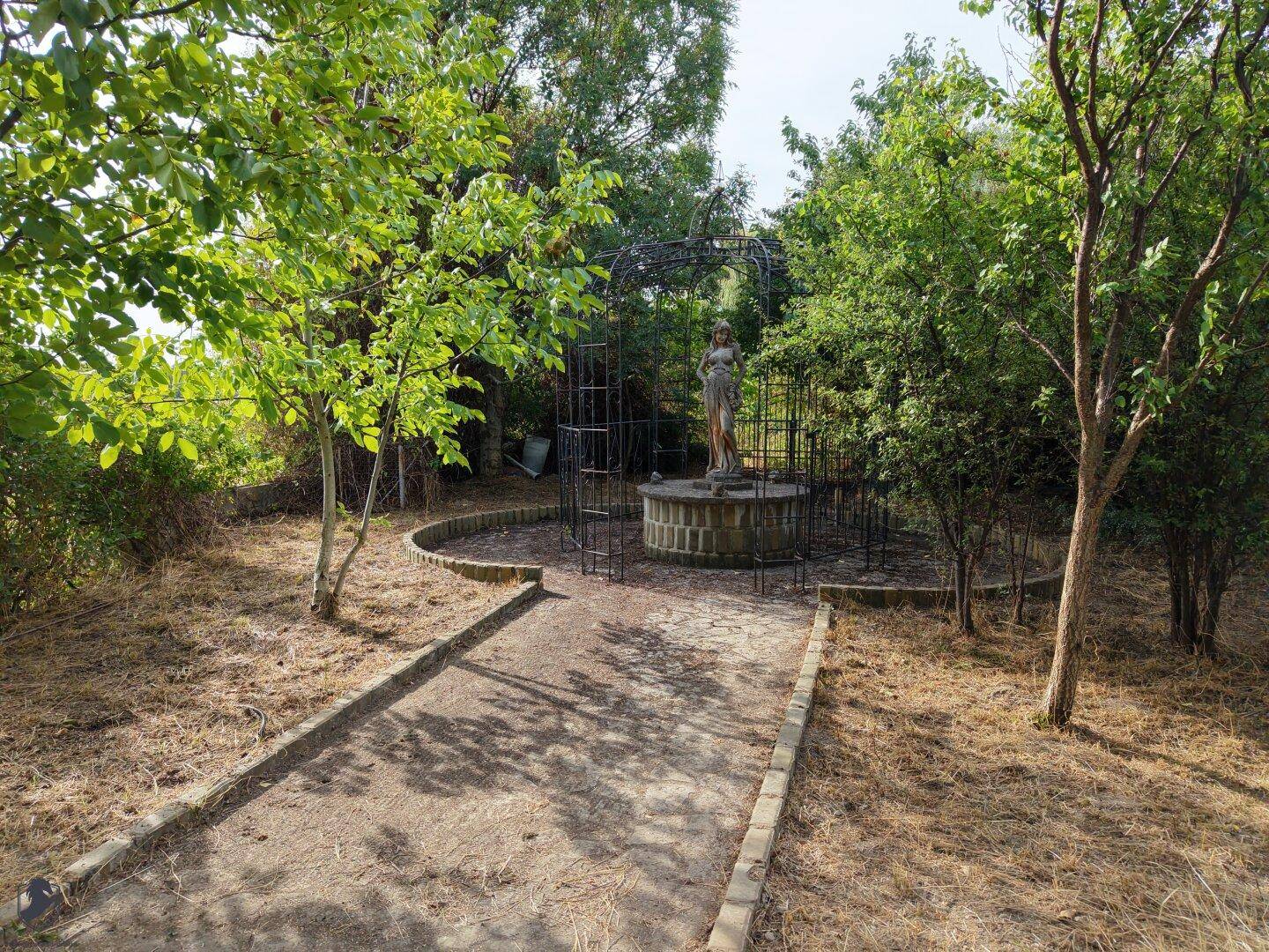 Gartenweg mit einer dekorativen Statue in einem Metallkäfig, umgeben von Bäumen und Sträuchern.