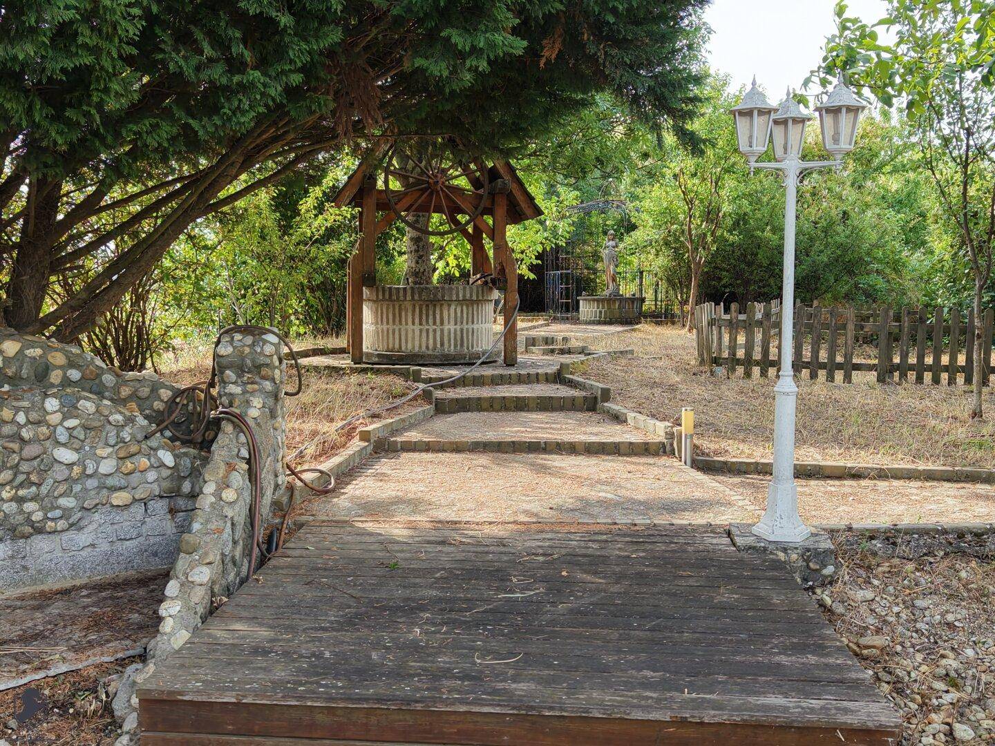Gartenweg mit einem dekorativen Holzbrunnen, Laternen und einer Holzbrücke.