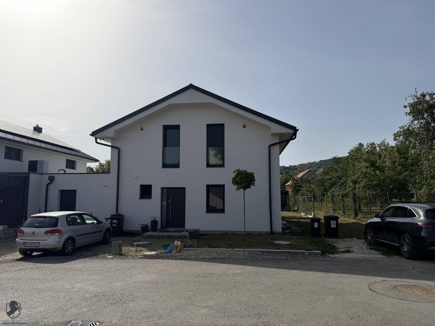 Modernes Einfamilienhaus mit weißer Fassade, dunklen Fenstern und Satteldach in sonniger Lage.