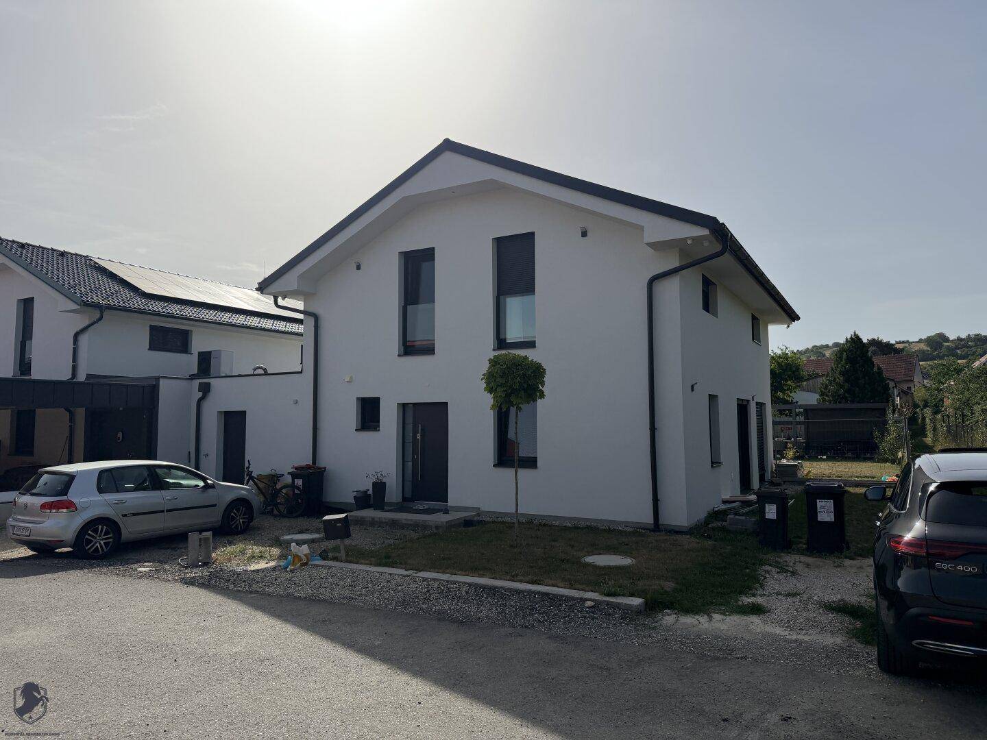 Neubau-Einfamilienhaus mit weißer Fassade, dunklen Fenstern und Vorgartenbereich an einem sonnigen Tag.