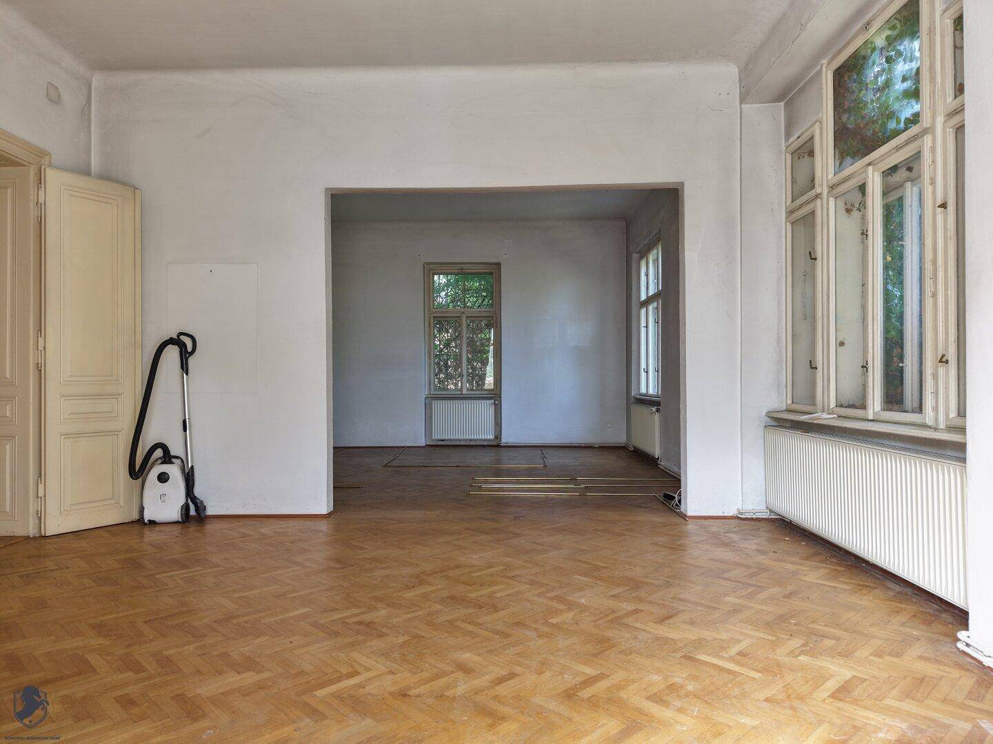 Geräumiges Wohnzimmer mit Fischgrätparkett, hohen Decken und großen Fenstern.