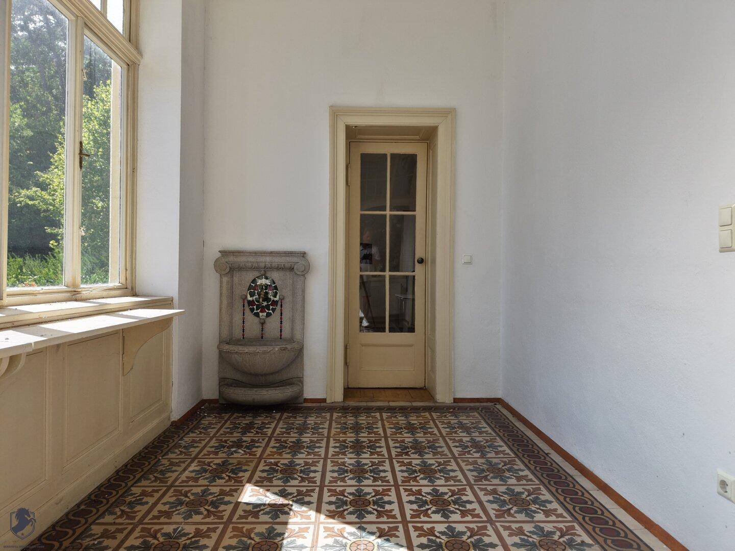 Heller Raum mit gemusterten Fliesen, einem dekorativen Wandbrunnen und großen Fenstern.