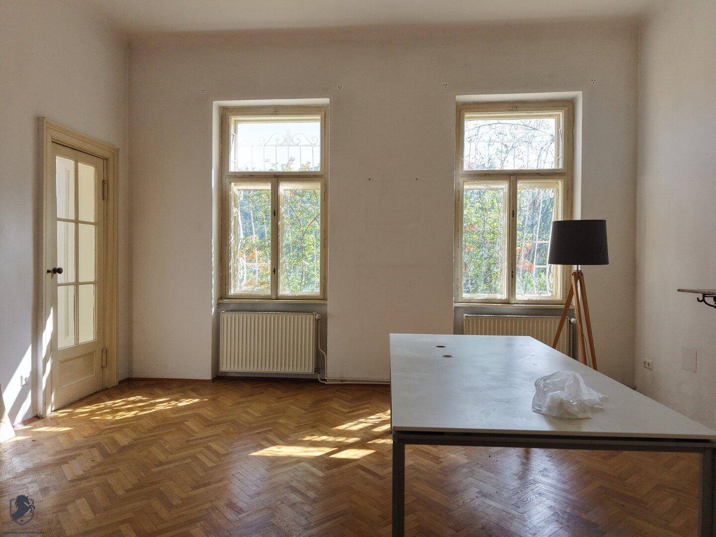 Helles Arbeitszimmer mit Fischgrätparkett, hohen Fenstern und einem großen Tisch.