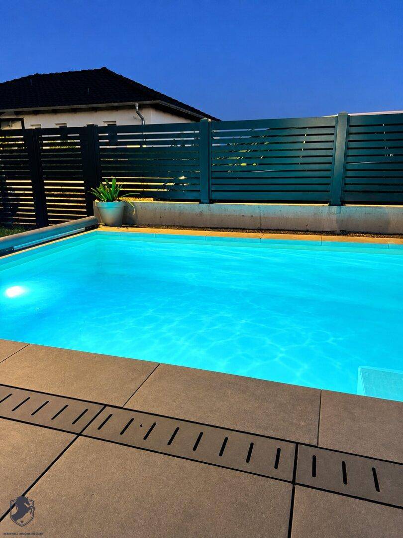 Beleuchteter Swimmingpool am Abend mit moderner Umrandung und Zaun.