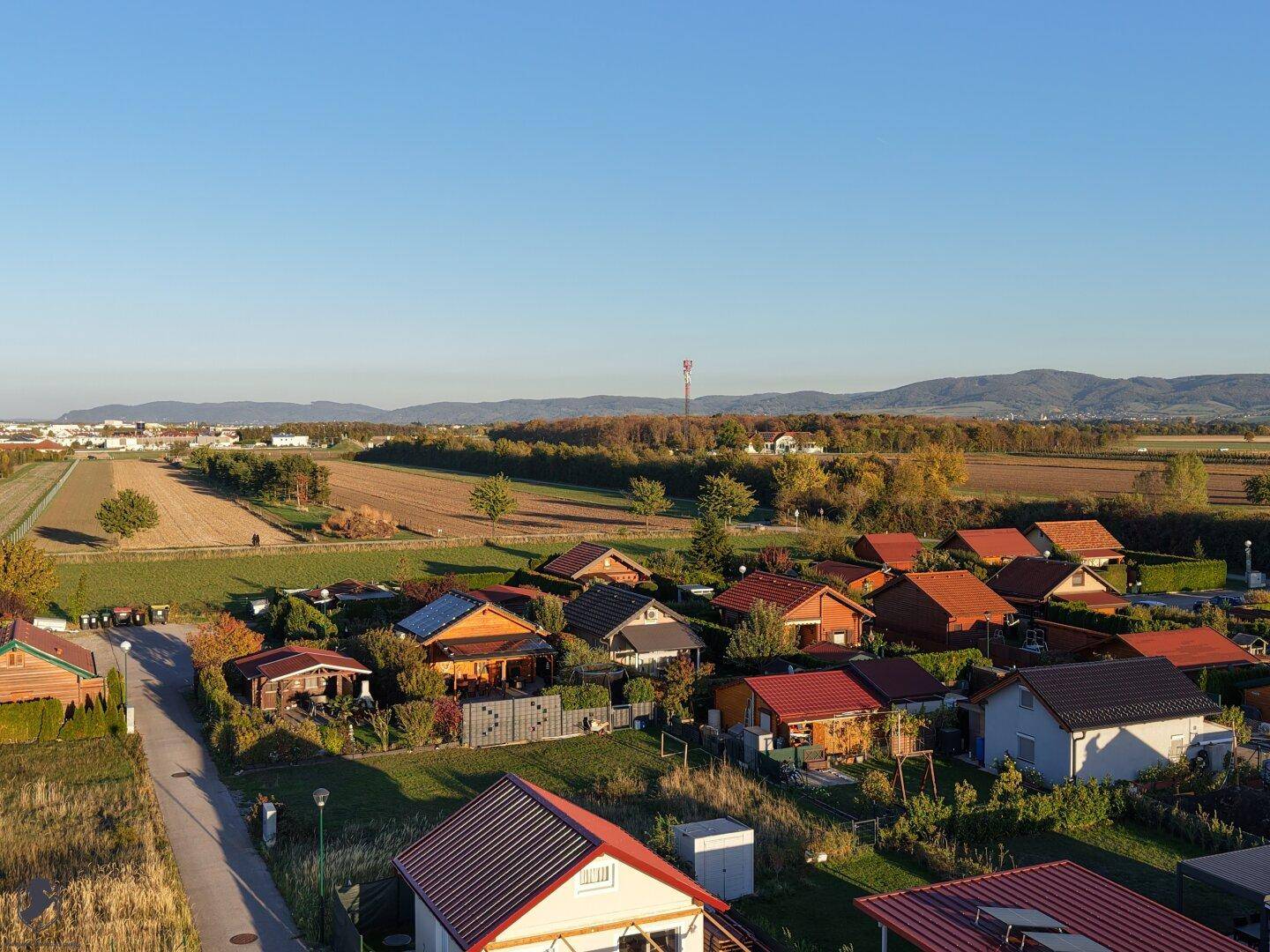 Weitwinkelaufnahme einer Wohnsiedlung umgeben von Feldern mit Bergpanorama am Horizont.
