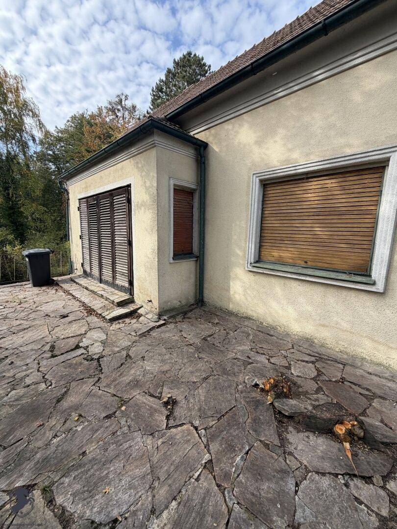Gepflasterter Außenbereich am Haus mit Fensterläden und einem Mülleimer.