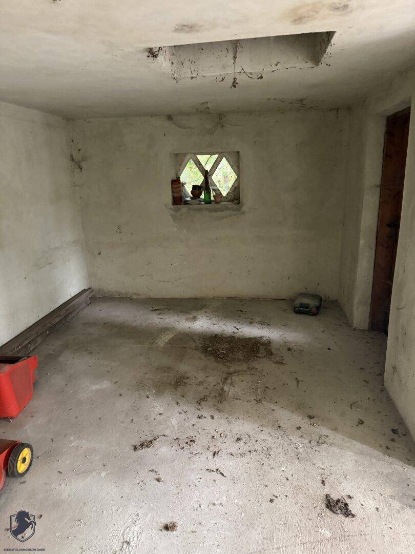 Einfacher, unfertiger Raum mit Betonboden und einem kleinen Fenster, möglicherweise ein Abstellraum.