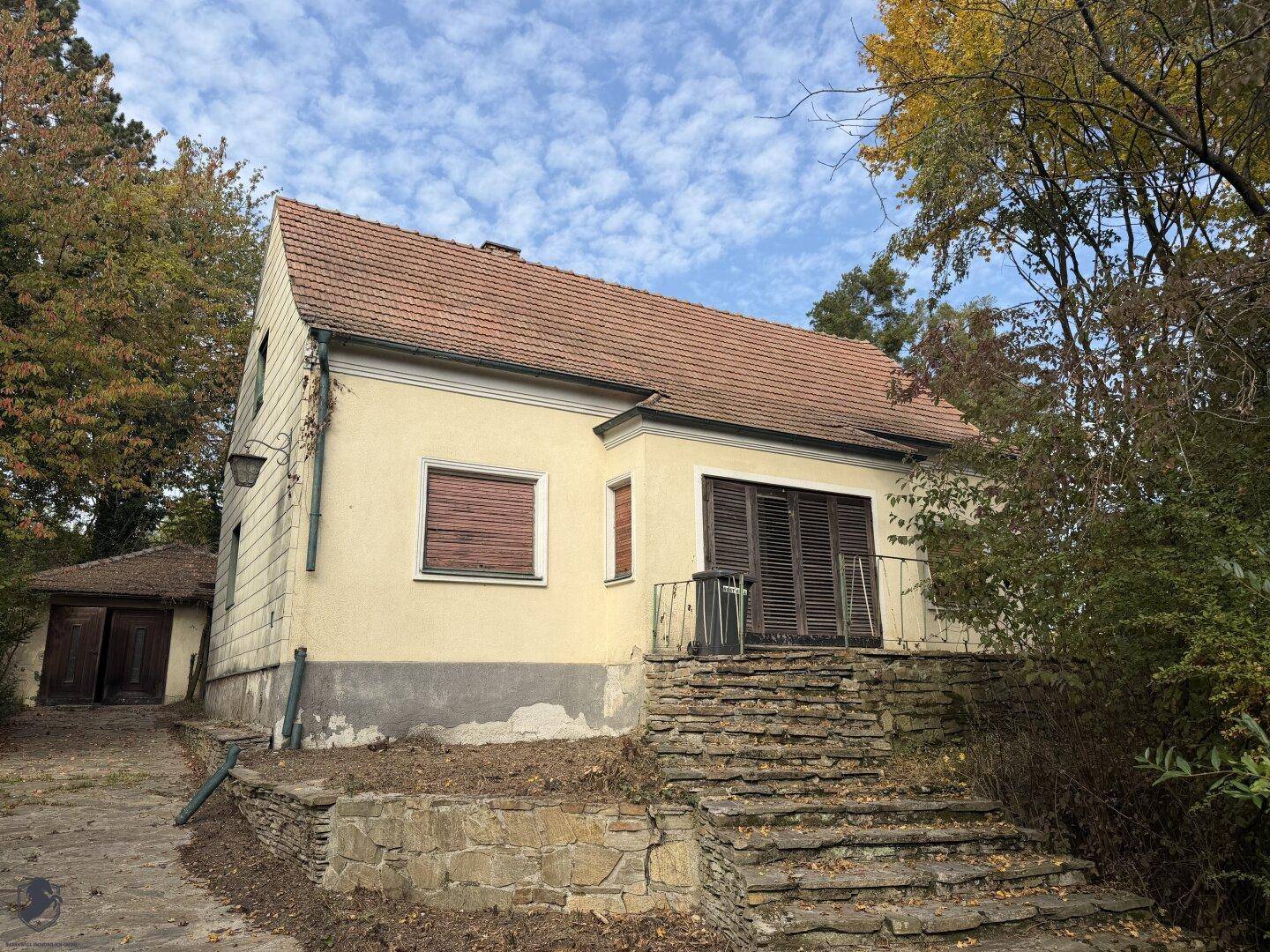 Älteres Wohnhaus mit Satteldach, Steintreppe und geschlossenen Fensterläden, umgeben von Bäumen.