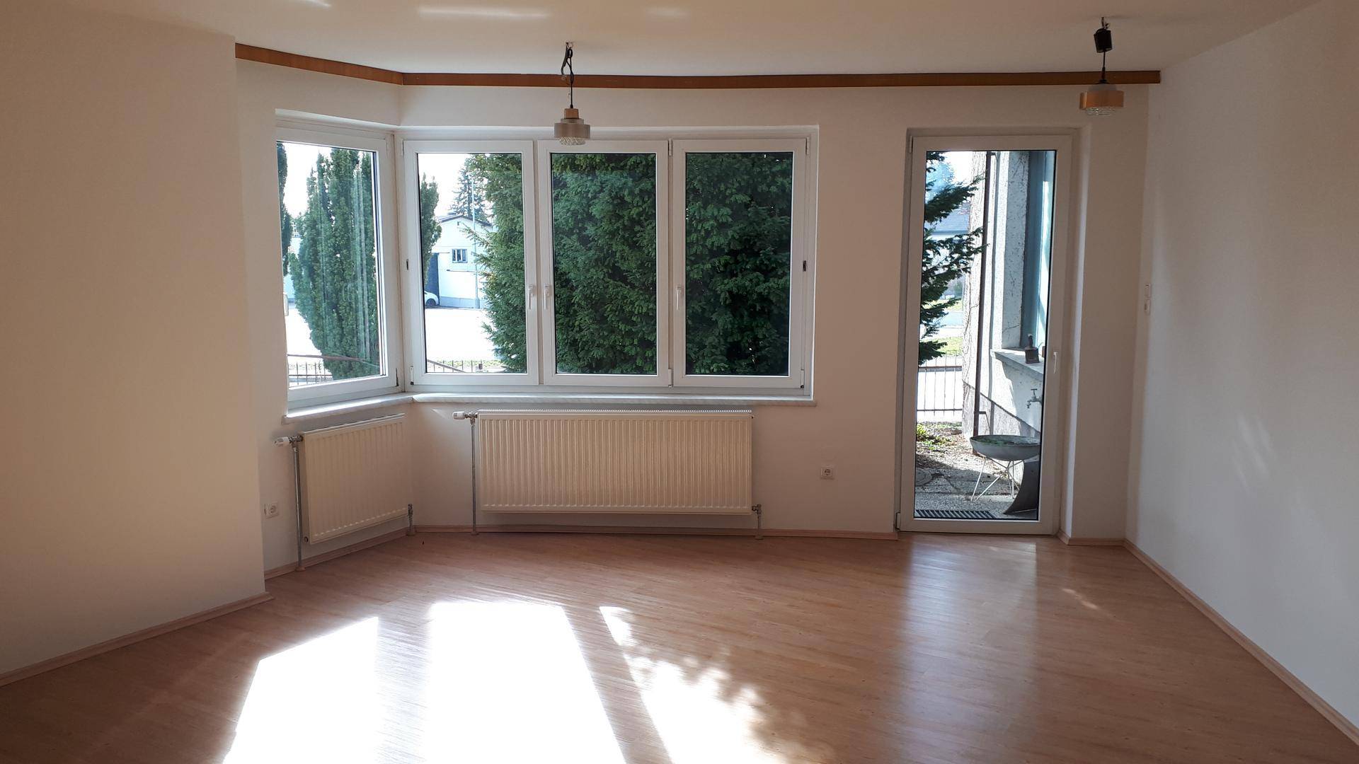 Lichtdurchfluteter Wohnraum mit großen Fenstern, Heizkörpern und Zugang zu einem Außenbereich.