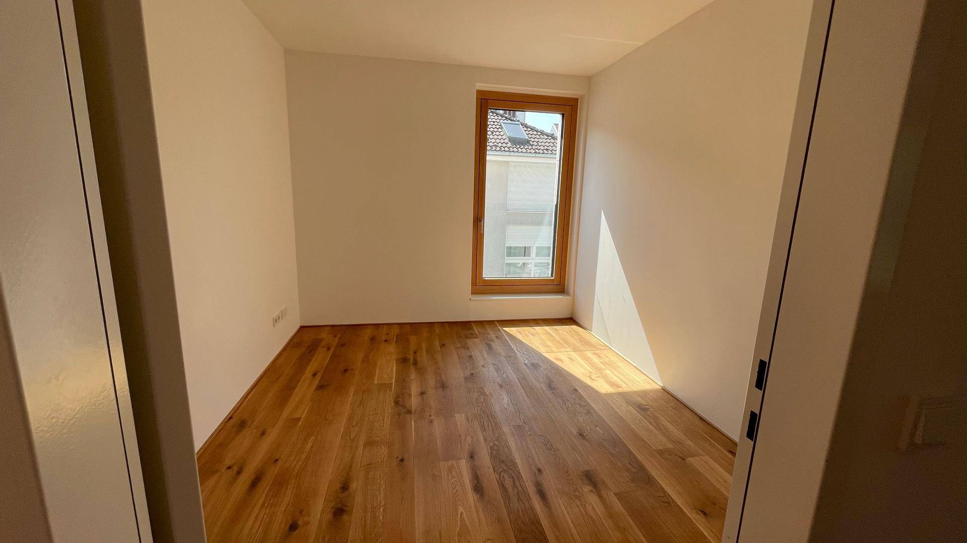 Helles Schlafzimmer mit Holzboden und einem hohen Fenster, das viel Licht spendet.