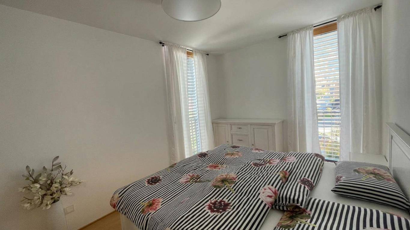Helles Schlafzimmer mit Doppelbett, floraler Bettwäsche und großen Fenstern mit Jalousien.