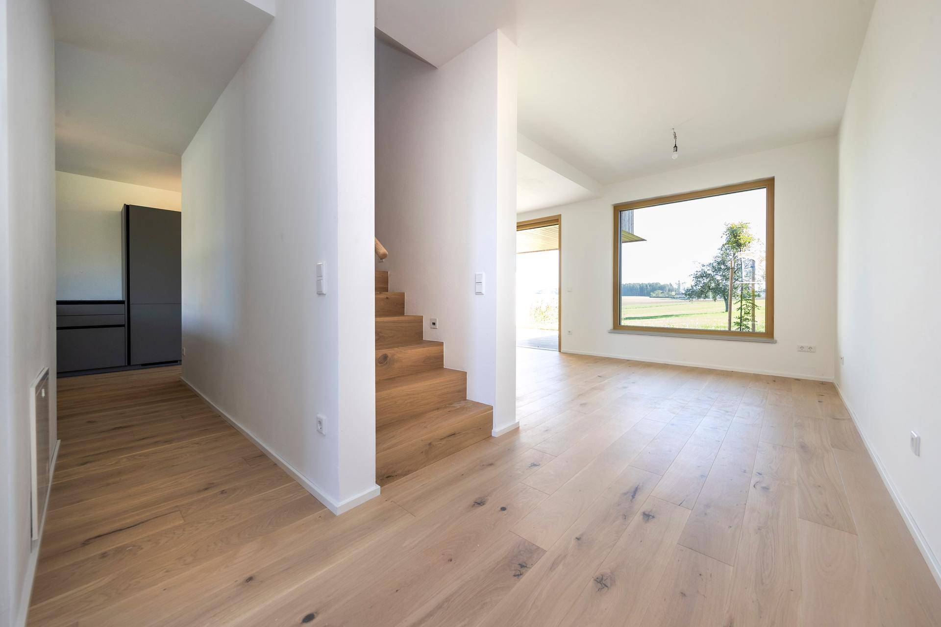 Offener Wohnbereich mit Holzboden, Treppenaufgang und großen Fenstern.