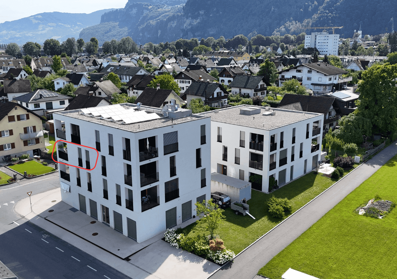 Moderne Wohngebäude mit Balkonen und Flachdächern, umgeben von einer grünen Landschaft und Bergen.