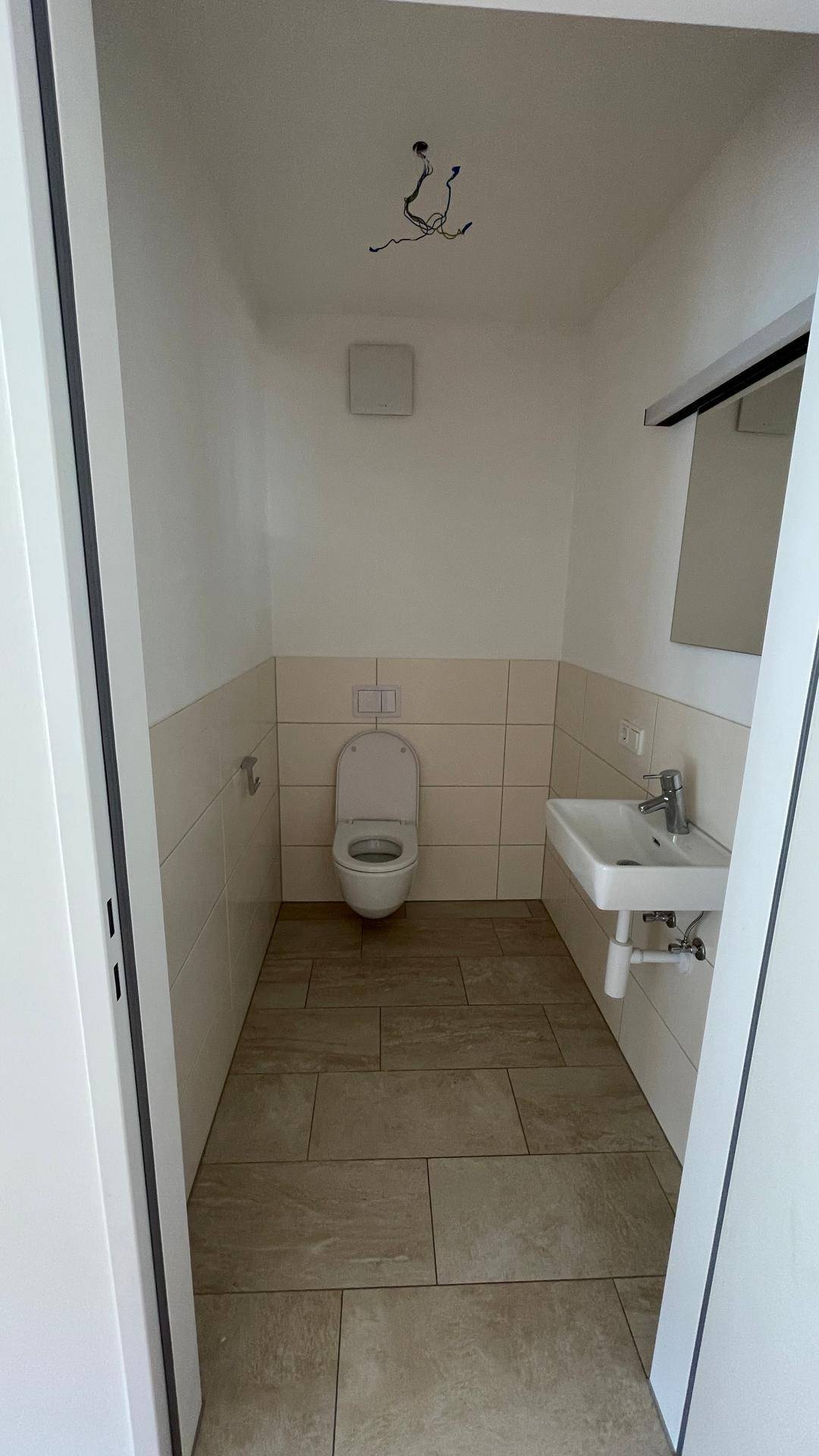 Gäste-WC mit Wand-WC, kleinem Waschbecken und hellen Fliesen an Wänden und Boden.