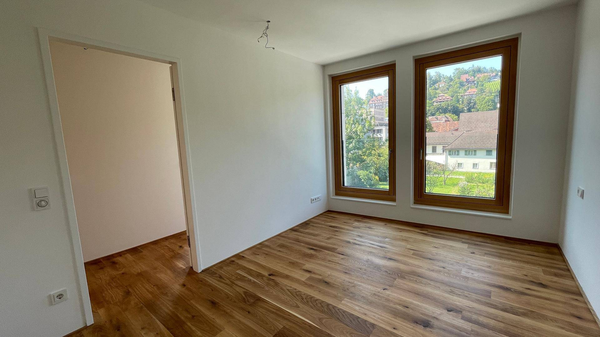 Schlafzimmer mit Holzboden und zwei großen Fenstern, die viel Tageslicht hereinlassen.