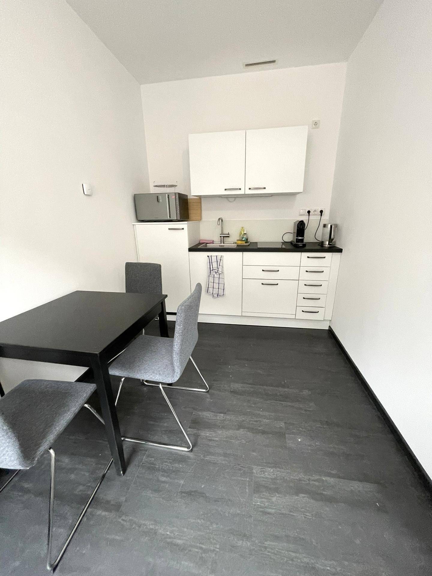 Kleine, moderne Küchenzeile mit weißen Schränken, Esstisch und dunklem Boden in einem hellen Raum.