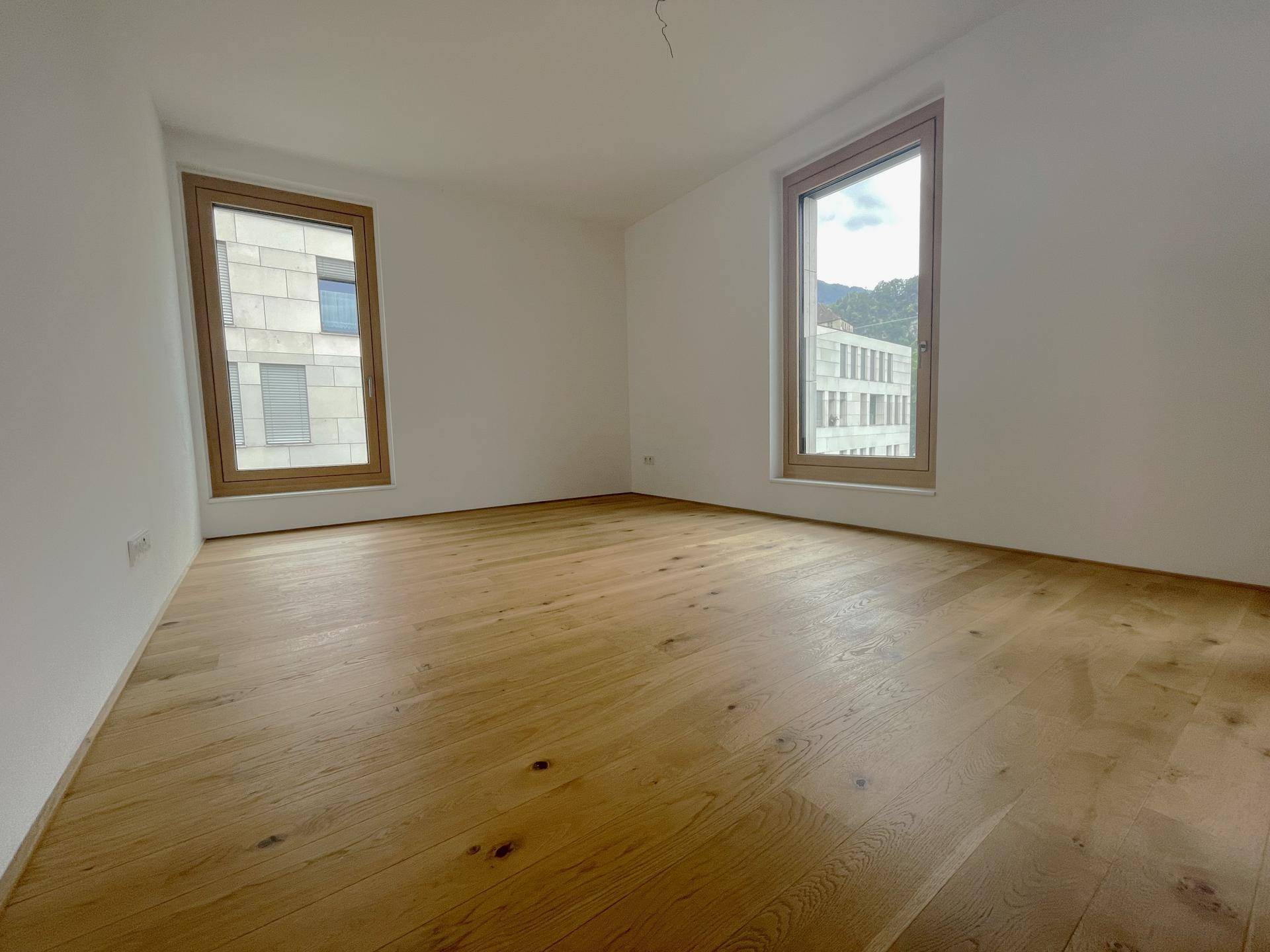 Heller, leerer Raum mit neuem Holzboden und zwei großen Fenstern.