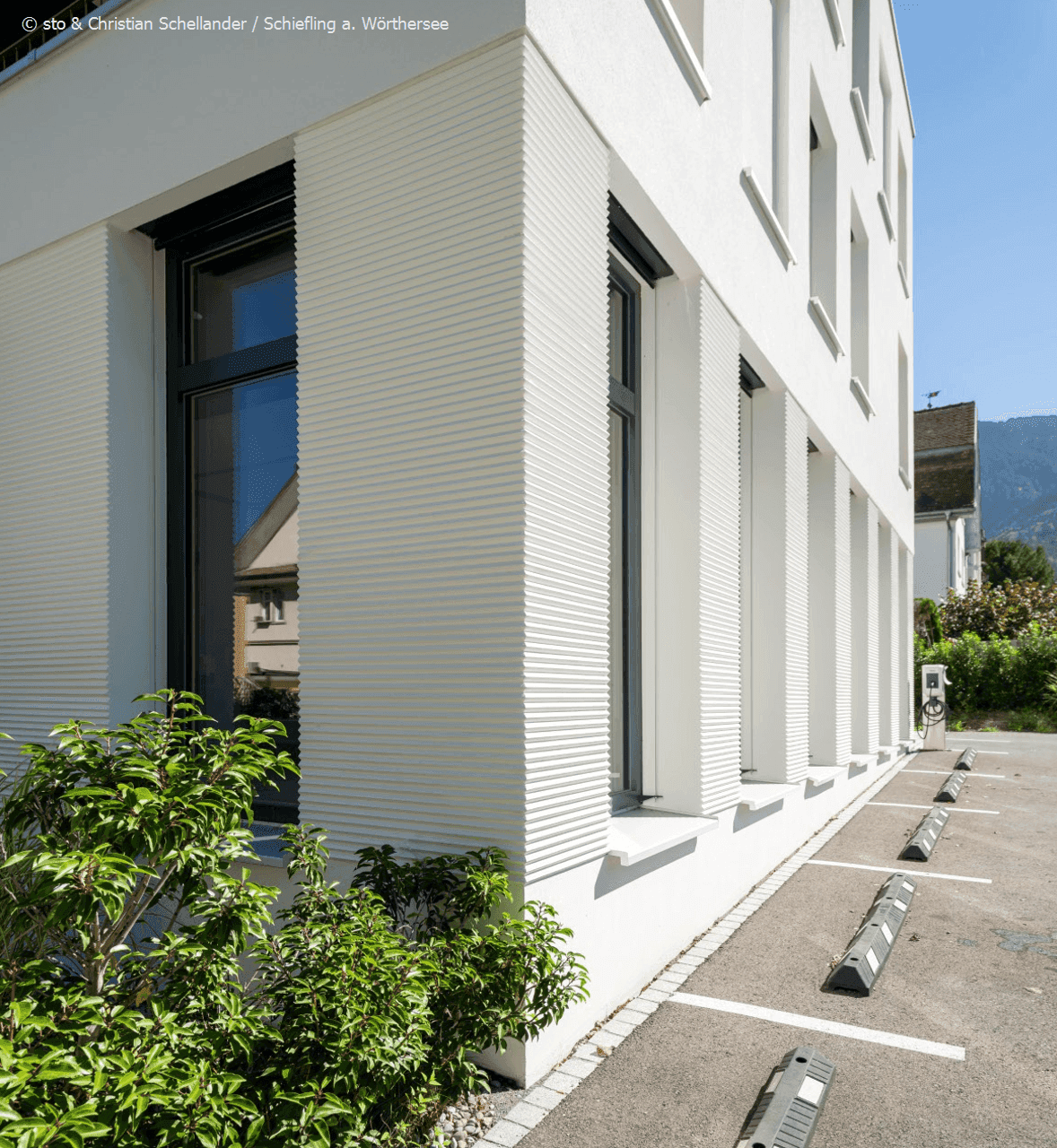 Nahaufnahme der modernen weißen Fassade mit strukturierter Oberfläche und großen Fenstern, inklusive Ladestation.