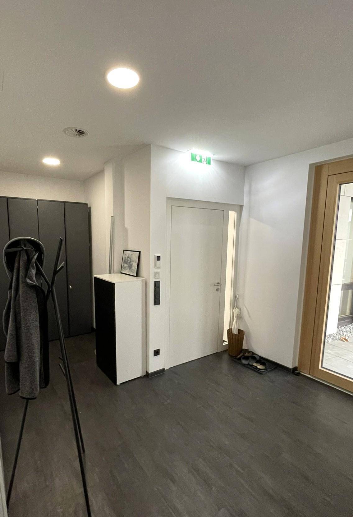 Heller Eingangsbereich mit dunklem Boden, Garderobe und einer modernen Holztür, die nach außen führt.