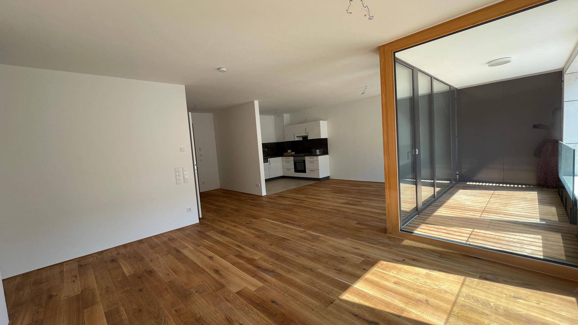 Offener Wohnbereich mit Holzboden, angrenzender Küche und Zugang zur verglasten Loggia.