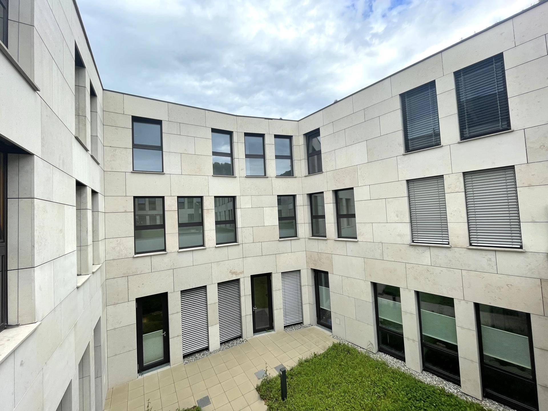 Blick in einen modernen Innenhof mit heller Steinfassade, großen Fenstern und einer kleinen Grünfläche.