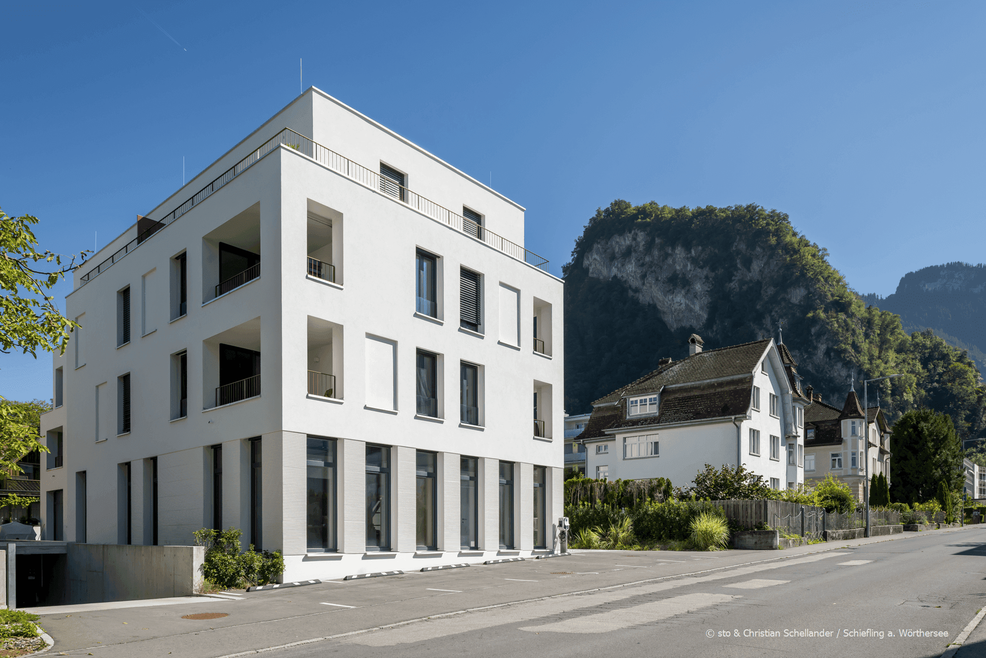 Modernes Gebäude in alpiner Umgebung mit Bergblick, umgeben von älteren Bauten und Parkplätzen.