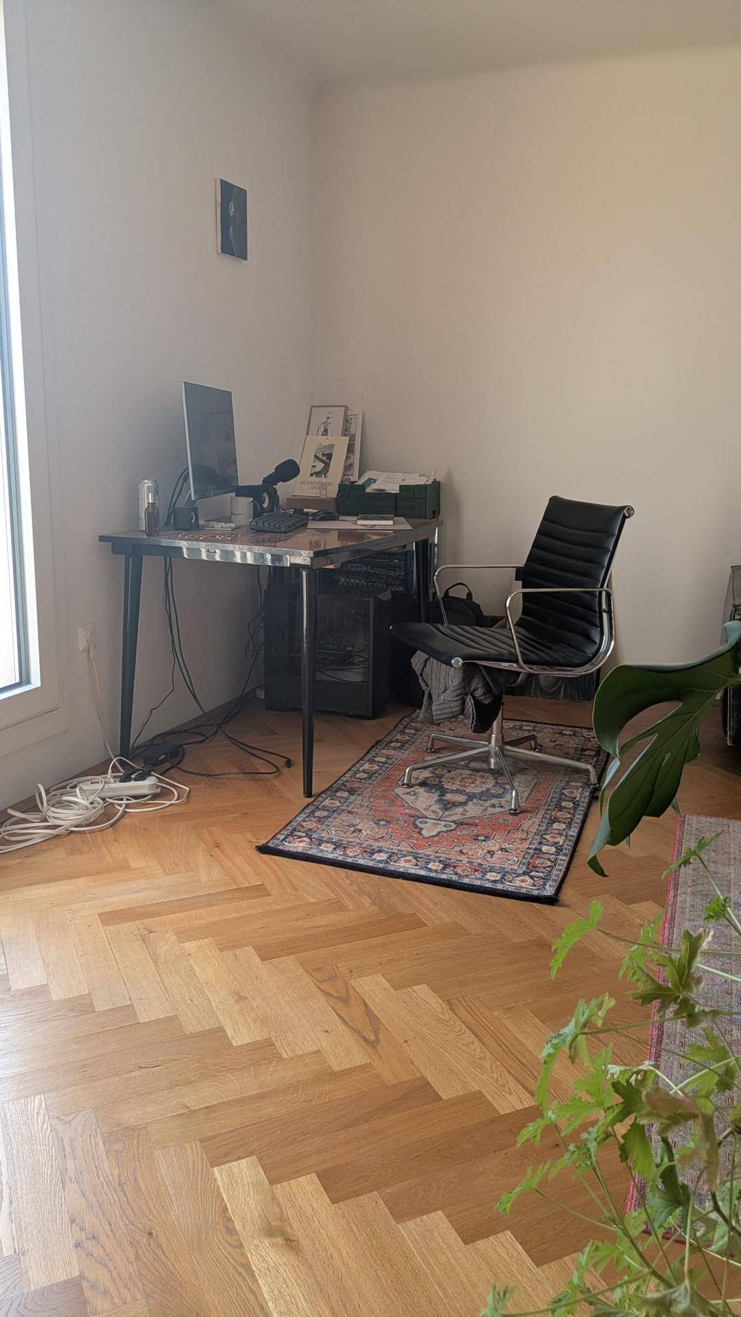 Arbeitszimmer mit Schreibtisch, Bürostuhl und Fischgrätparkett, gut beleuchtet.