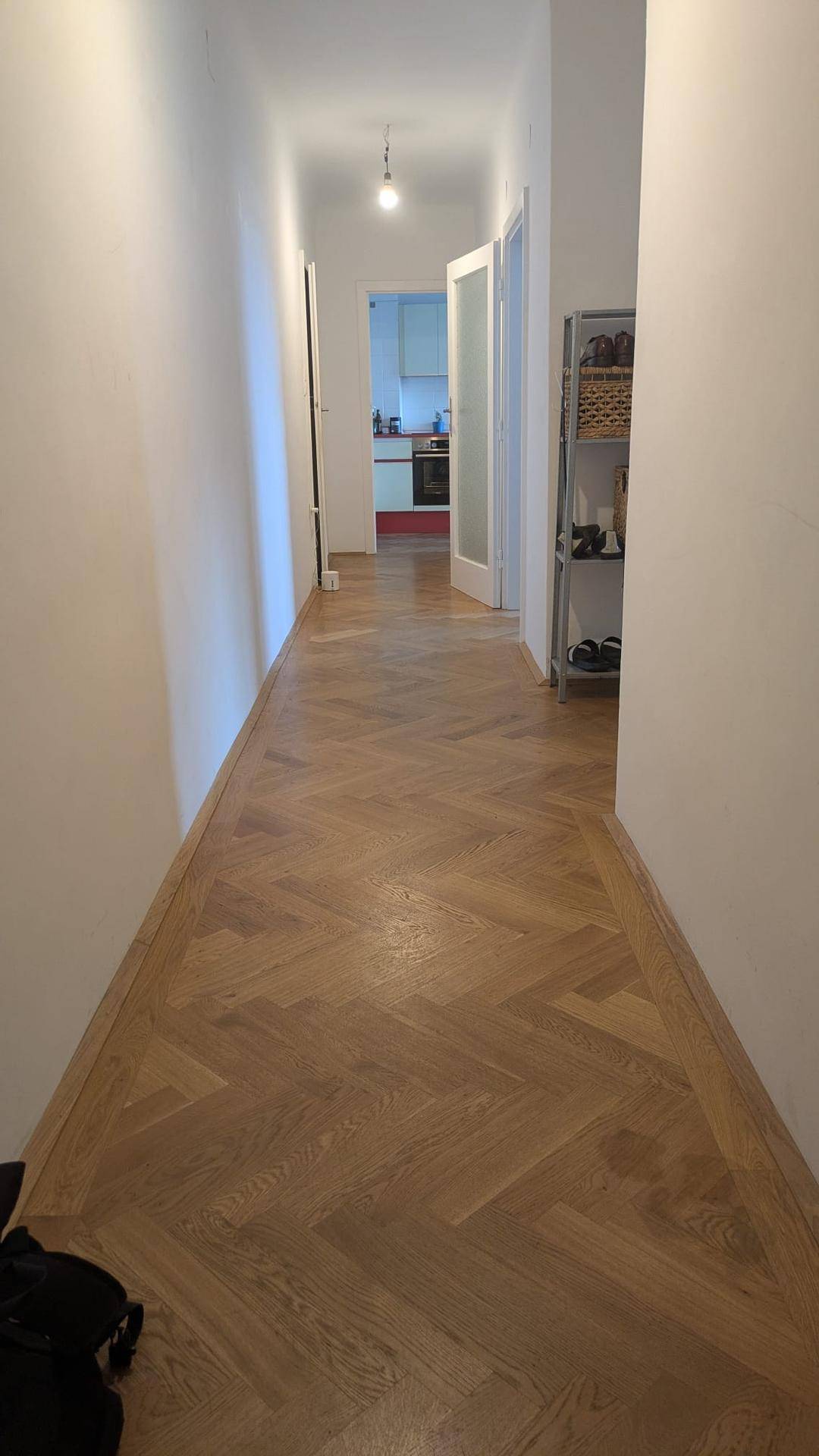 Langer Flur mit hellem Fischgrätparkett, weißen Wänden und Blick in die Küche.