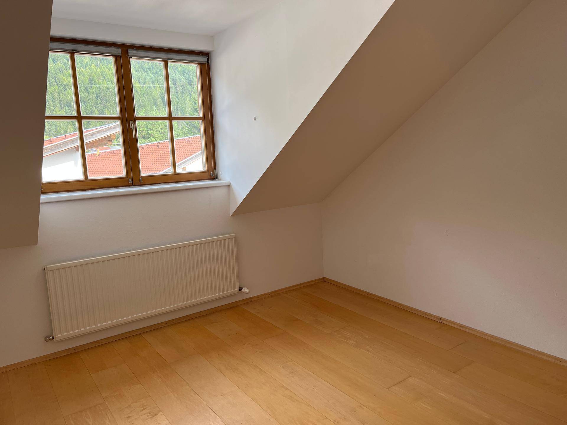 Gemütliches Zimmer im Dachgeschoss mit hellem Holzparkett und Fenster, bietet Blick ins Grüne.