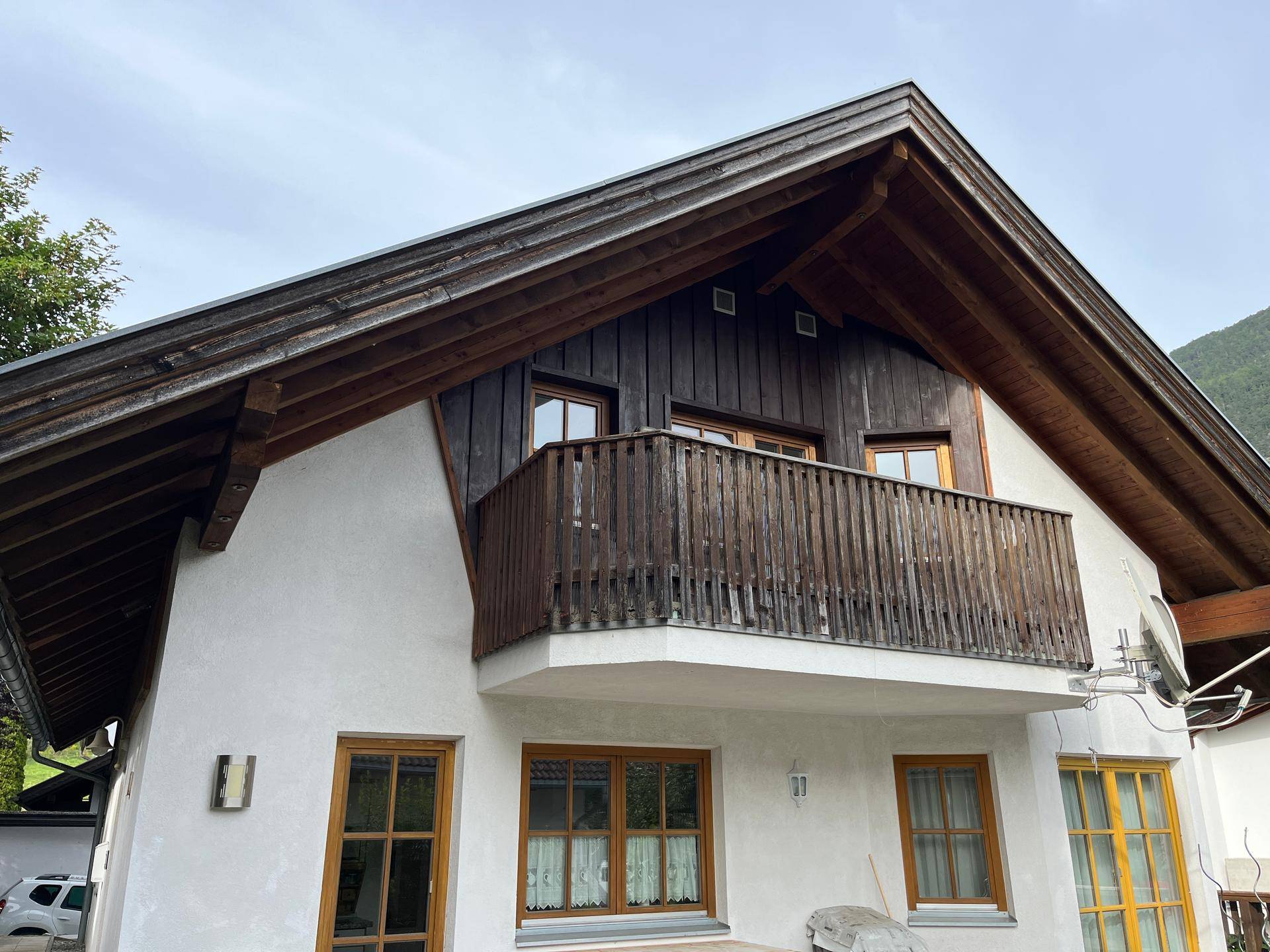 Vorderansicht des Hauses mit traditionellem Holzbalkon und Satteldach, umgeben von Grün.