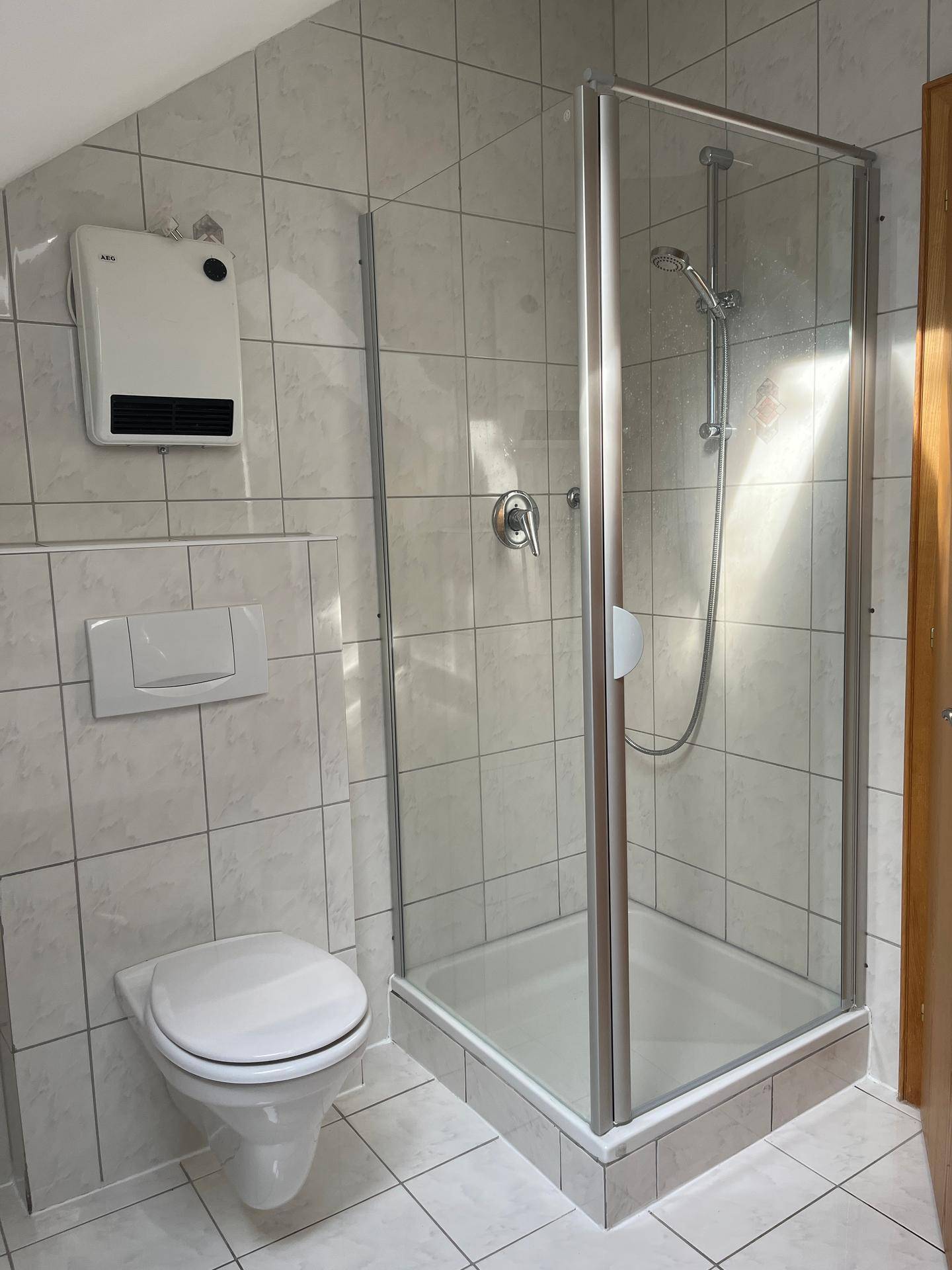 Gefliestes Badezimmer mit wandmontierter Toilette und Duschkabine, ausgestattet mit einem Heizlüfter.