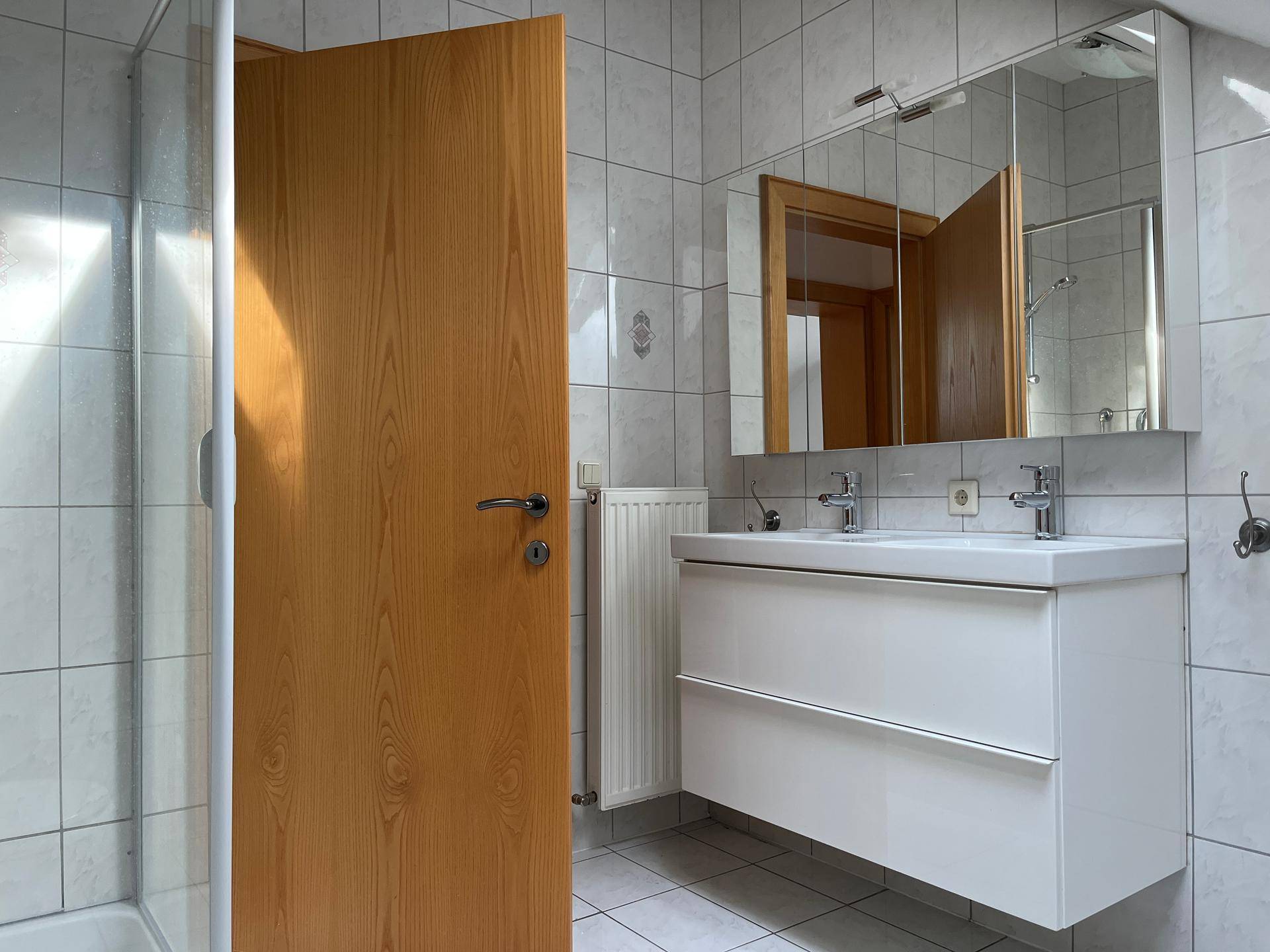 Modernes Badezimmer mit Doppelwaschbecken, Spiegelschrank und Duschkabine, hell gefliest.