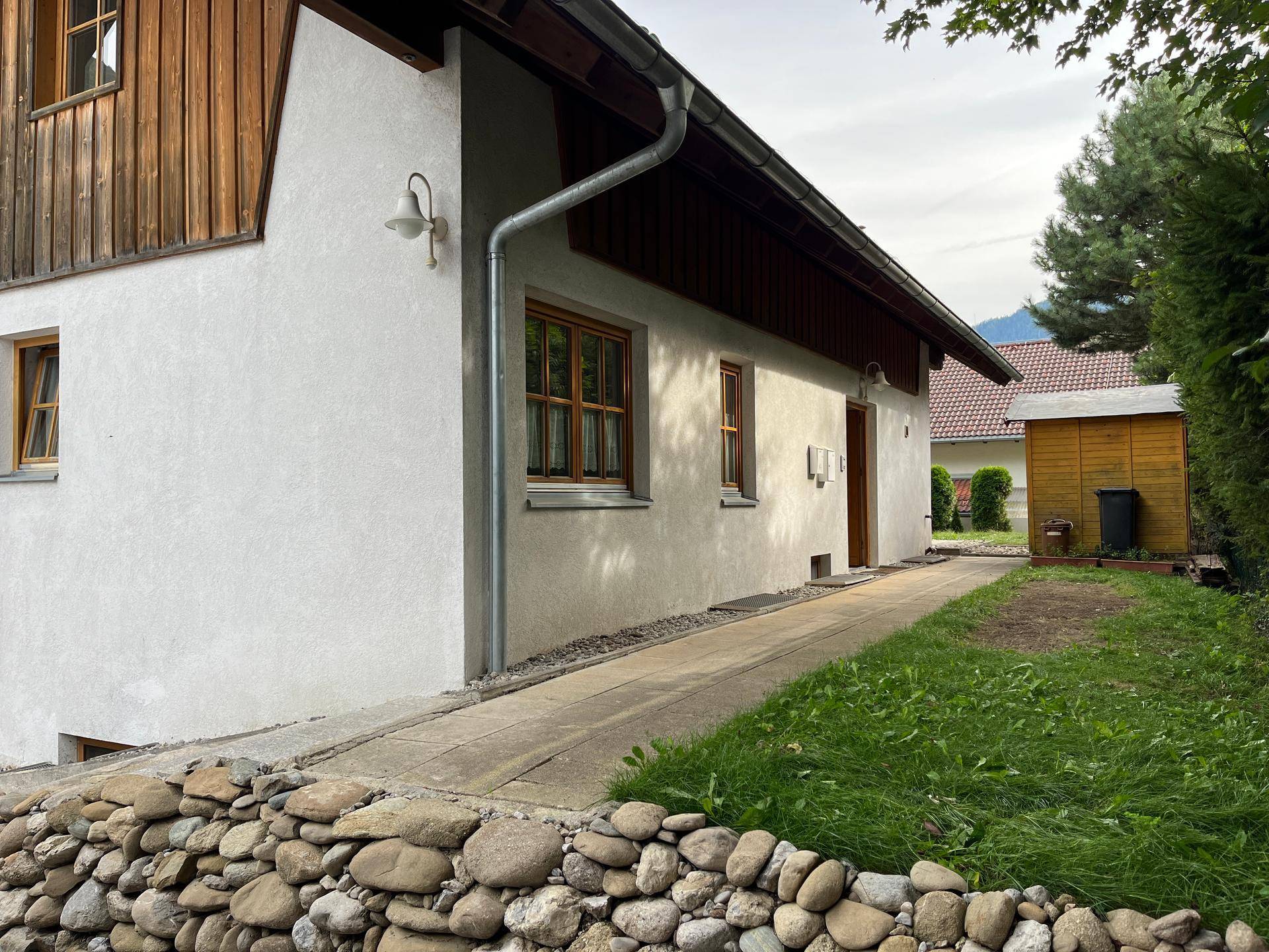 Seitenansicht des Hauses mit weißer Fassade, Rampe und kleinem Gartenbereich mit Steinmauer.
