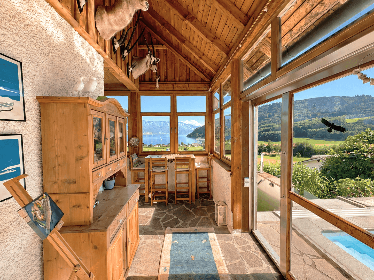 Heller Wintergarten mit Holzbalkendecke, Steinboden und Panoramablick auf den See und die Berge.