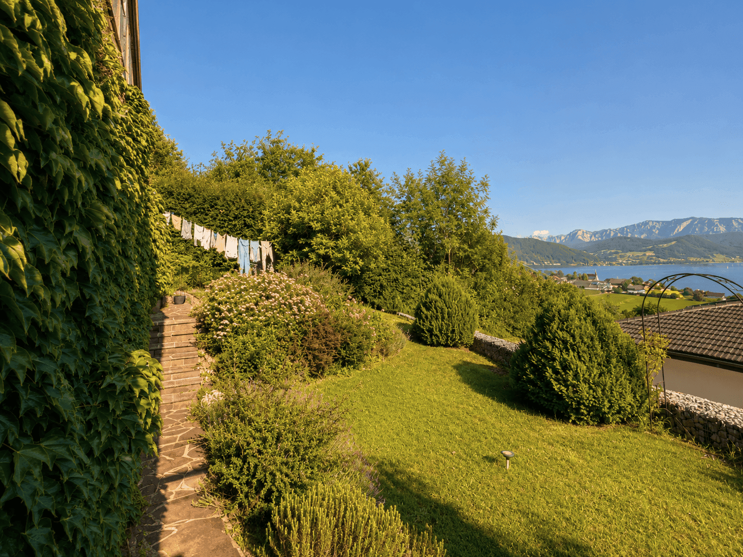Gepflegter Garten mit Steintreppe, Wäscheleine und weitem Blick über den See und die umliegende Landschaft.